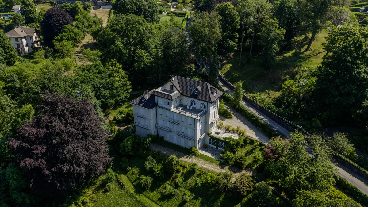 Aerial view of a multi-story white villa with a dark roof, surrounded by lush green trees and a winding driveway.
