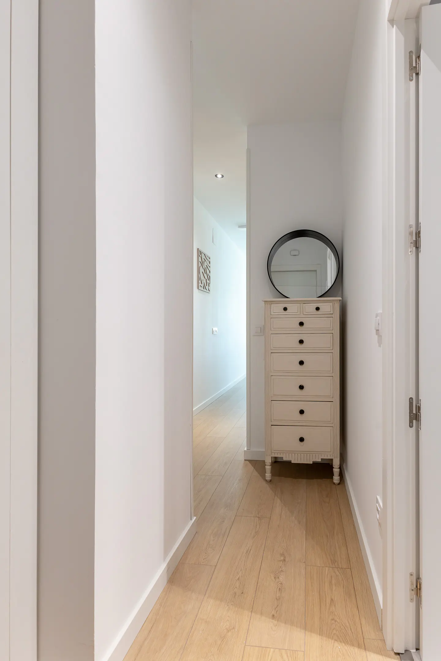 Bright hallway with light wood floors, white walls, and a white dresser with a black oval mirror on top.