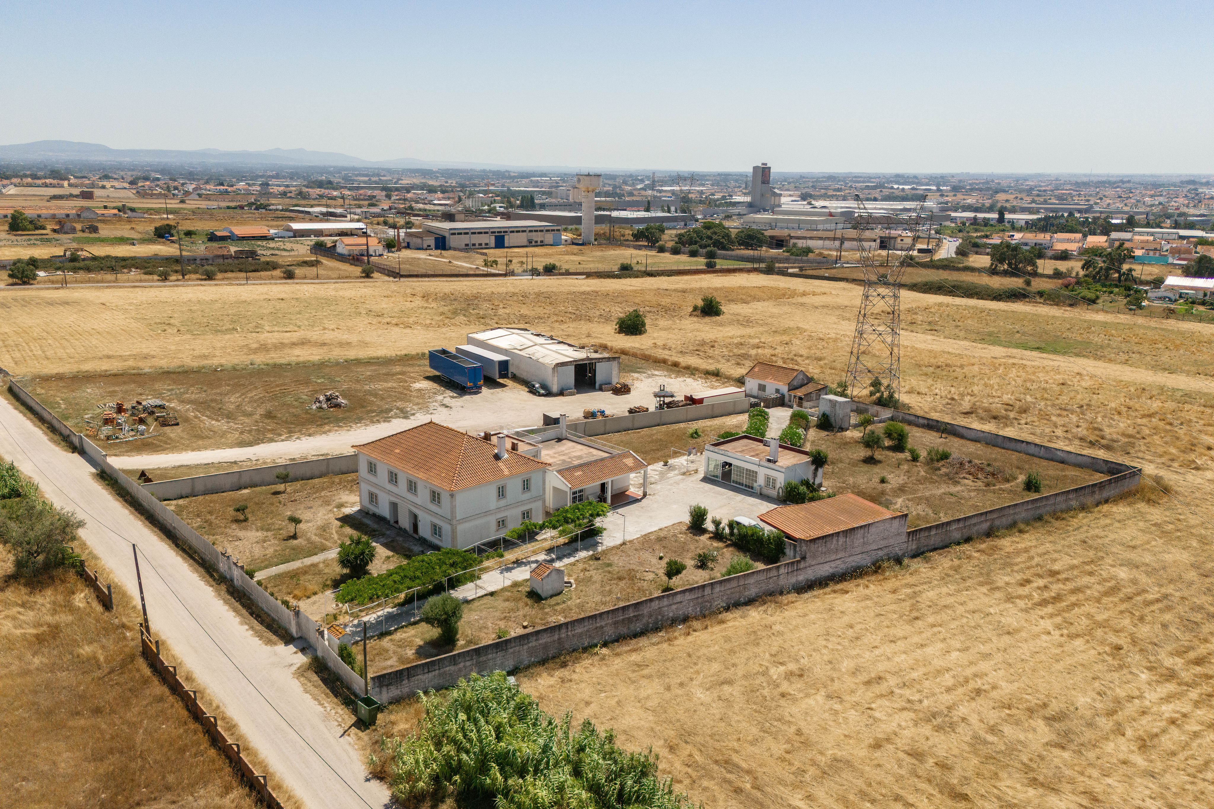 Aerial view of a walled property with a two-story house, outbuildings, and dry fields in a rural industrial area.