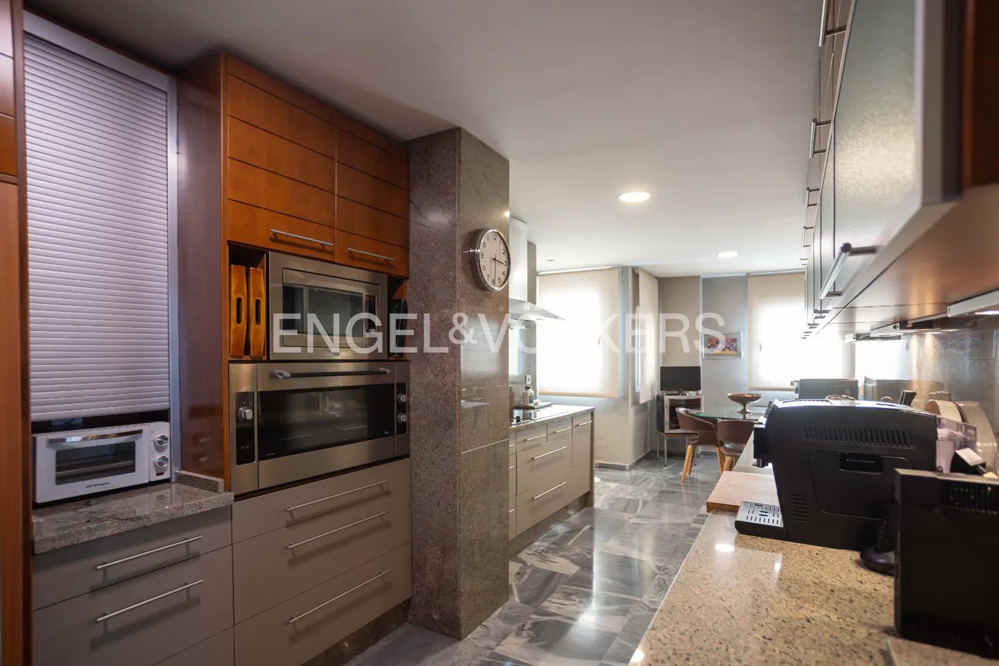 A modern kitchen with gray cabinets, stainless steel appliances, and a marble floor. A dining area is visible in the background.
