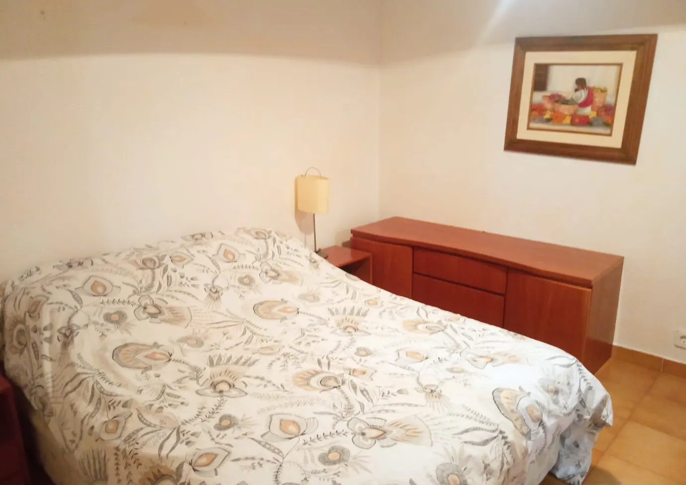Bedroom with a bed featuring a floral patterned comforter, a wooden dresser, a bedside lamp, and framed wall art.