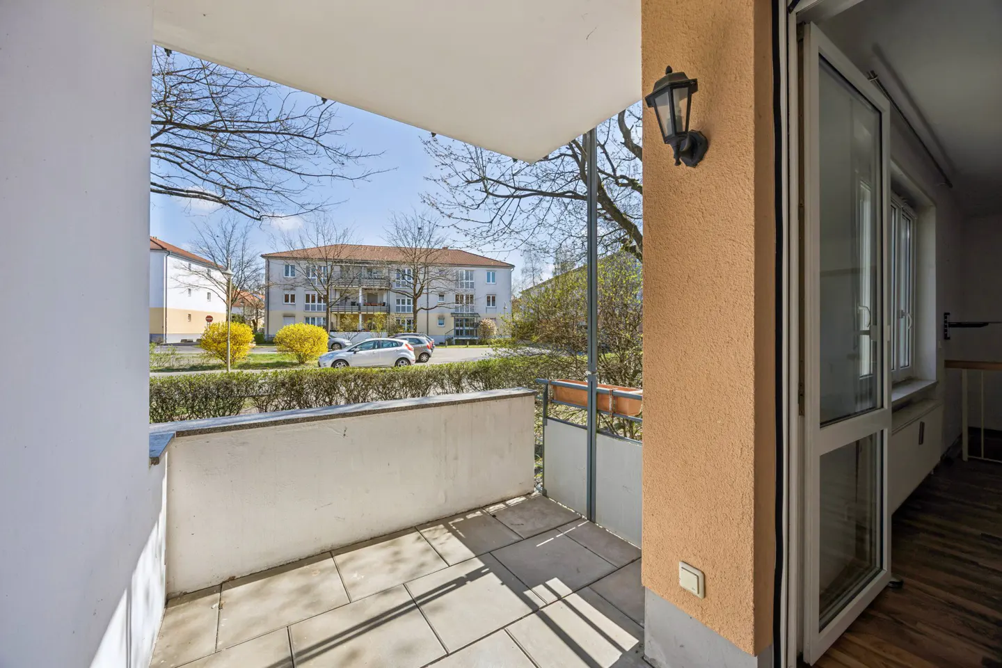 Balcony view with stone tiles, white walls, and an orange pillar with a black lantern. Outside, there are trees, bushes, cars, and a building under a blue sky.