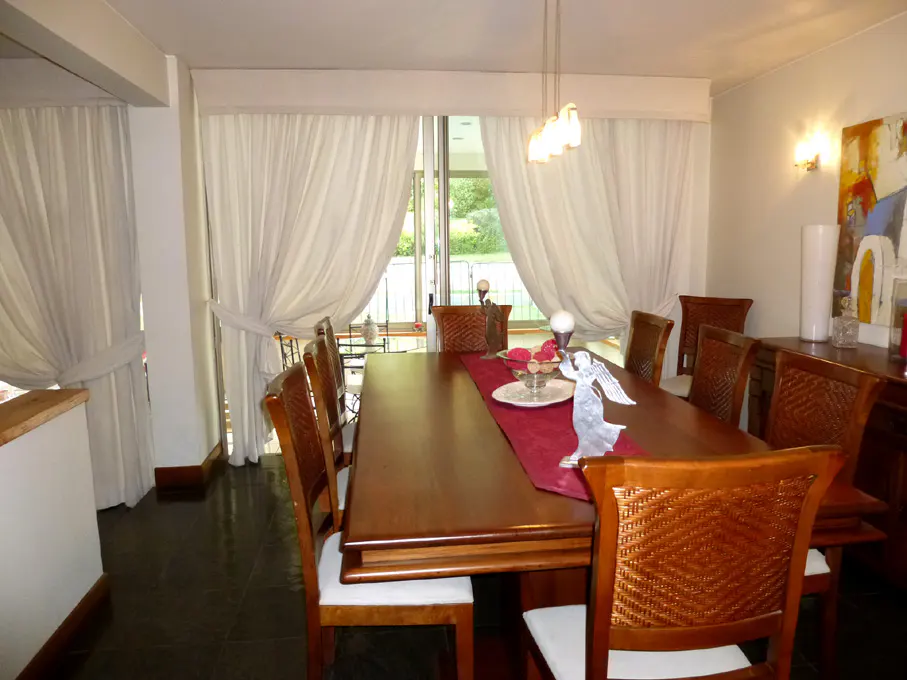 A dining room with a long wooden table, chairs, and white curtains. A red runner and decorations adorn the table.