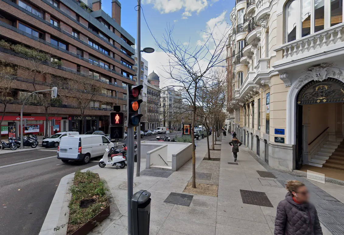 A city street scene with a red pedestrian traffic light, a white van, bare trees, and people walking on the sidewalk.
