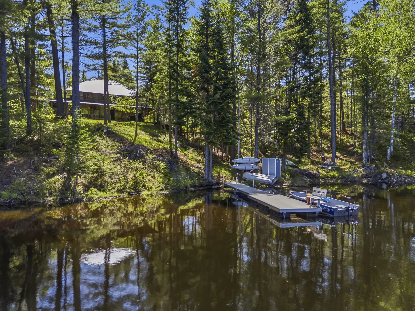 A house nestled in a lush, green forest sits above a calm lake with a private dock, small boat, and kayaks.