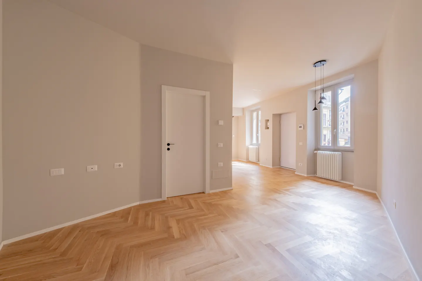 Bright, empty room with herringbone wood floors, white walls, and a white door. A window with a radiator is visible in the background.