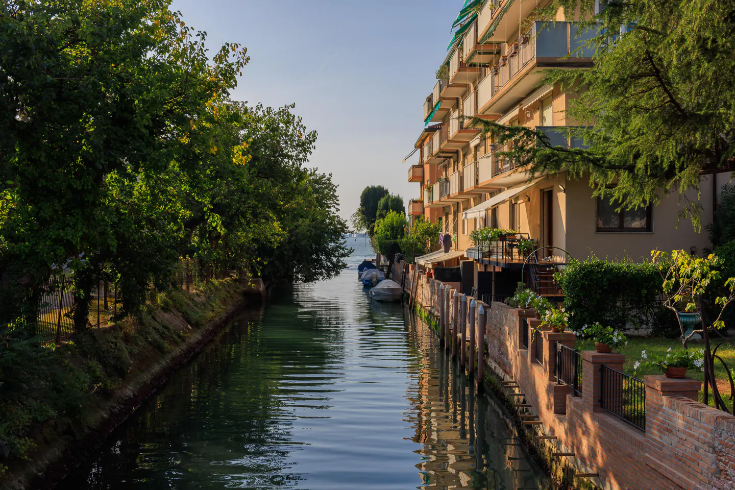 A canal runs between green trees and a building with balconies. Boats are docked in the distance.
