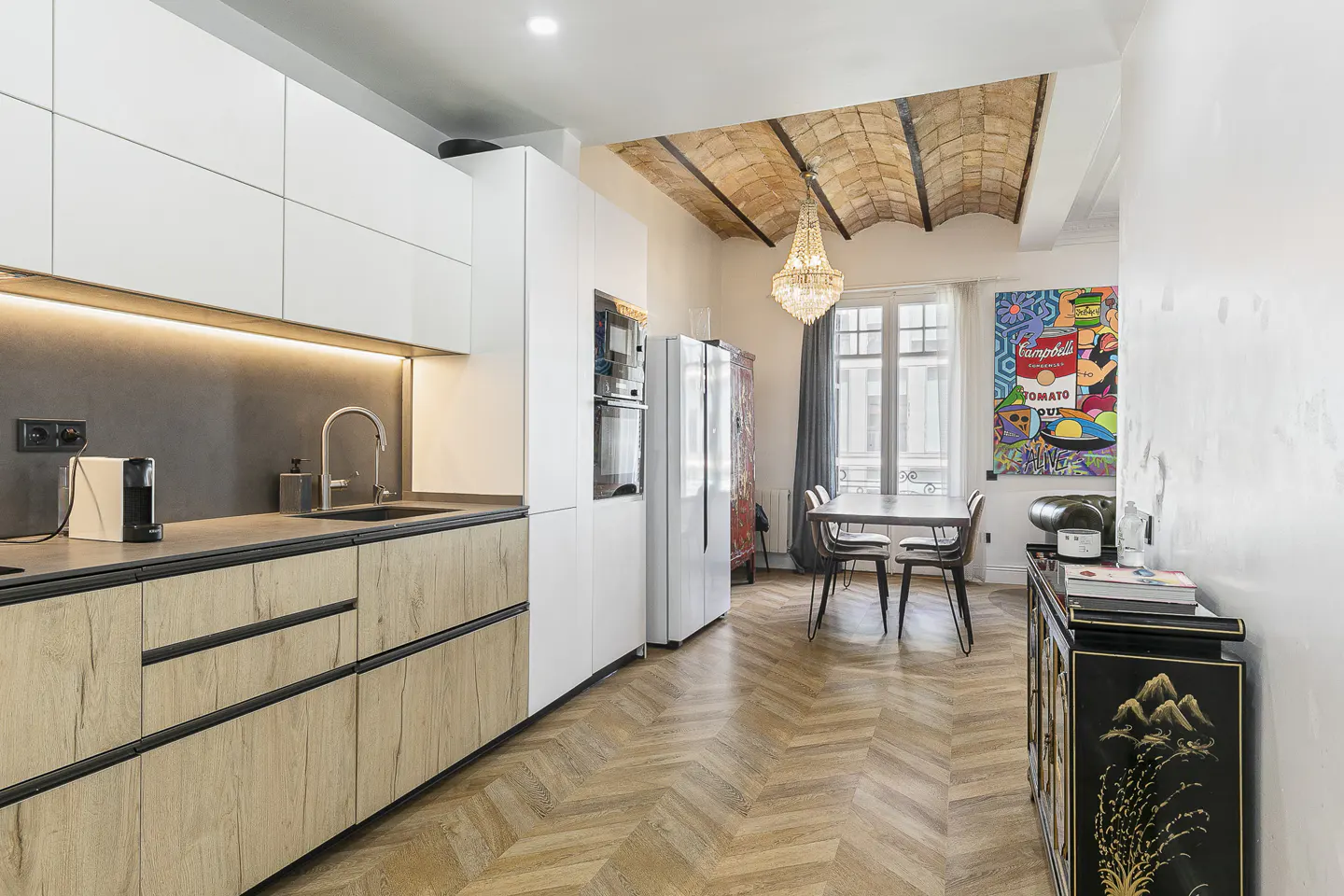 A modern kitchen with white cabinets, wood drawers, and herringbone floors. A dining table sits near a window, with a colorful pop art painting on the wall.