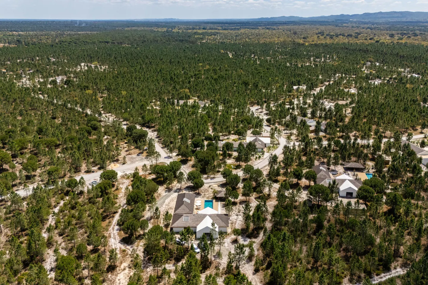Aerial view of white houses with pools nestled in a dense green forest under a bright blue sky.