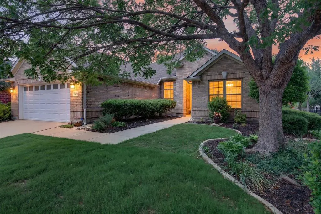 Brick house with a white garage door, green lawn, and a large tree at sunset.
