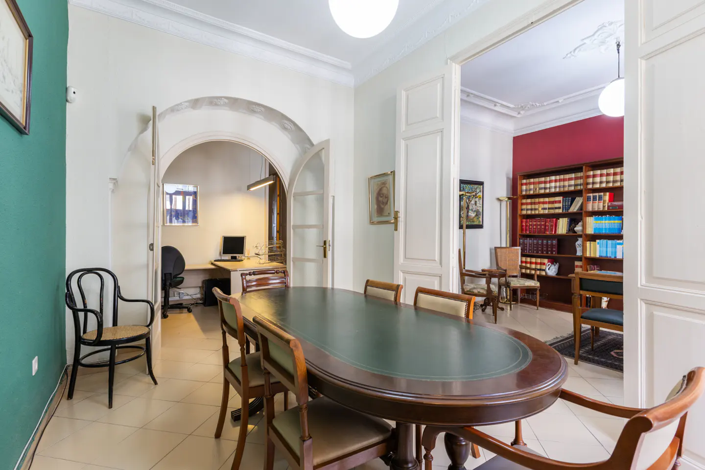 Interior view of a room with a large oval table, chairs, and arched doorways leading to an office and a library.