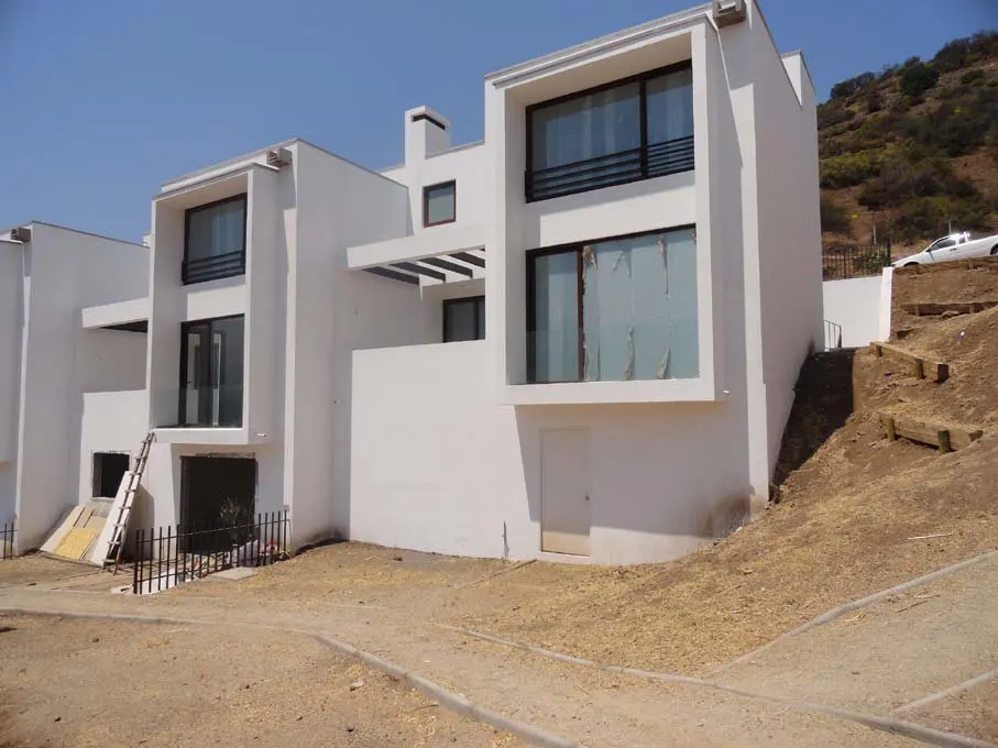 Modern white townhouses with large windows sit on a hillside under a blue sky. A dirt path leads to the homes.