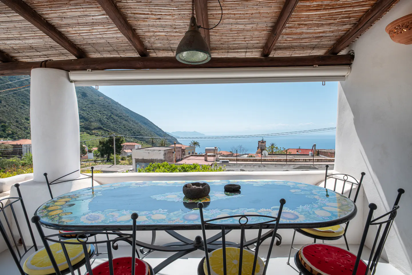 Outdoor dining area with a blue floral table, black chairs with colorful cushions, and a view of the sea and mountains.