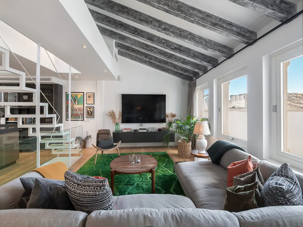 Bright living room with a gray sofa, green rug, and white staircase. A large TV hangs above a dark cabinet, and windows offer a view of the outside.