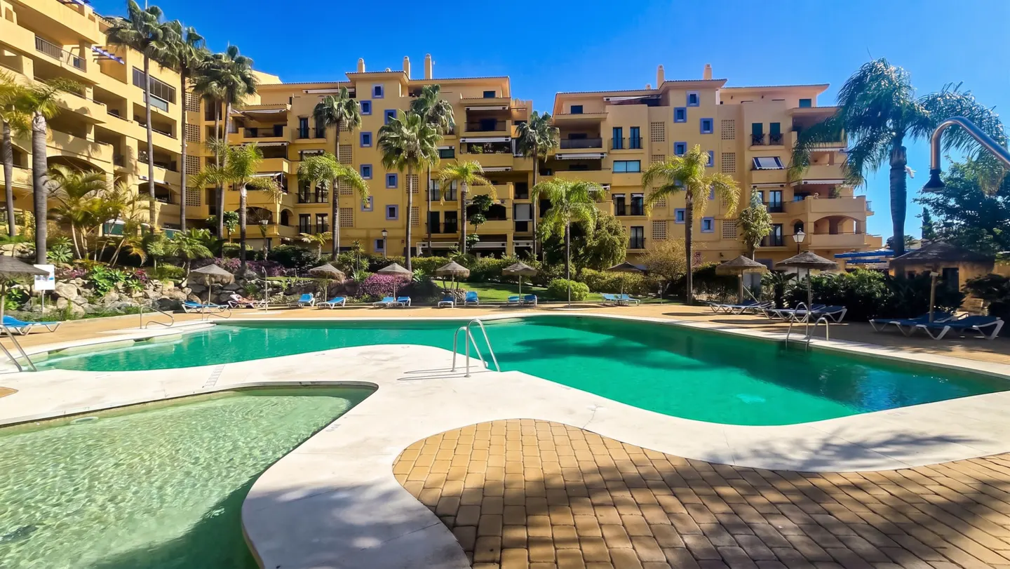 Large turquoise swimming pool in front of a multi-story yellow apartment complex with palm trees and lounge chairs.