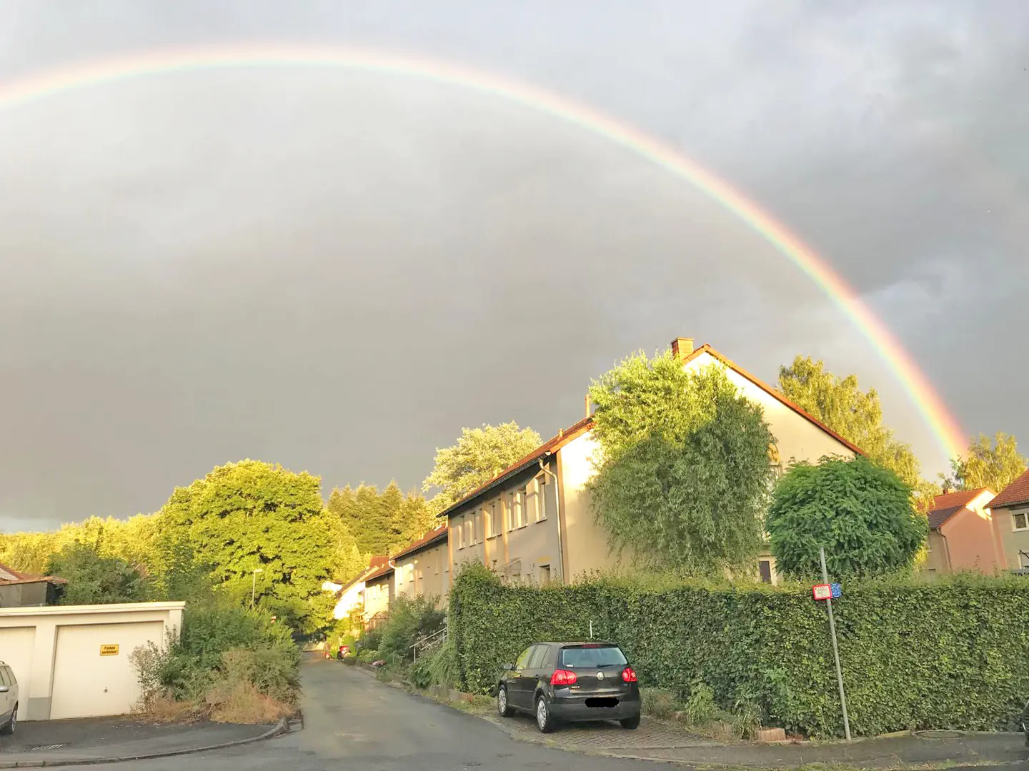 A rainbow arches over a street with houses, trees, and a parked black car.