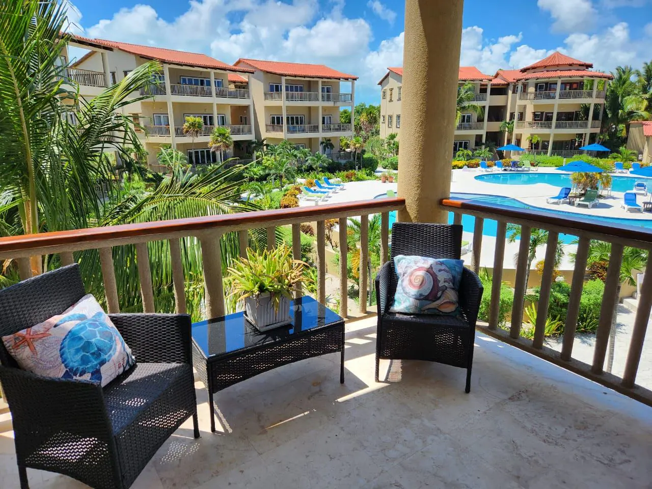 Balcony view of a resort with a pool. Two black chairs with pillows and a glass table are on the balcony. Palm trees and buildings are in the background.