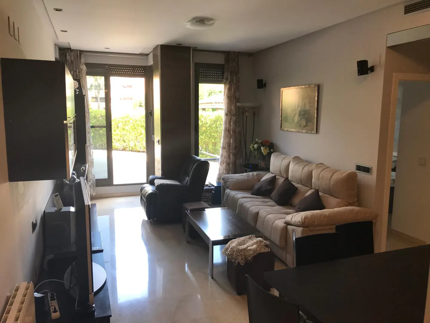 Bright living room with beige sofa, dark wood furniture, and a black leather recliner near a sliding glass door.