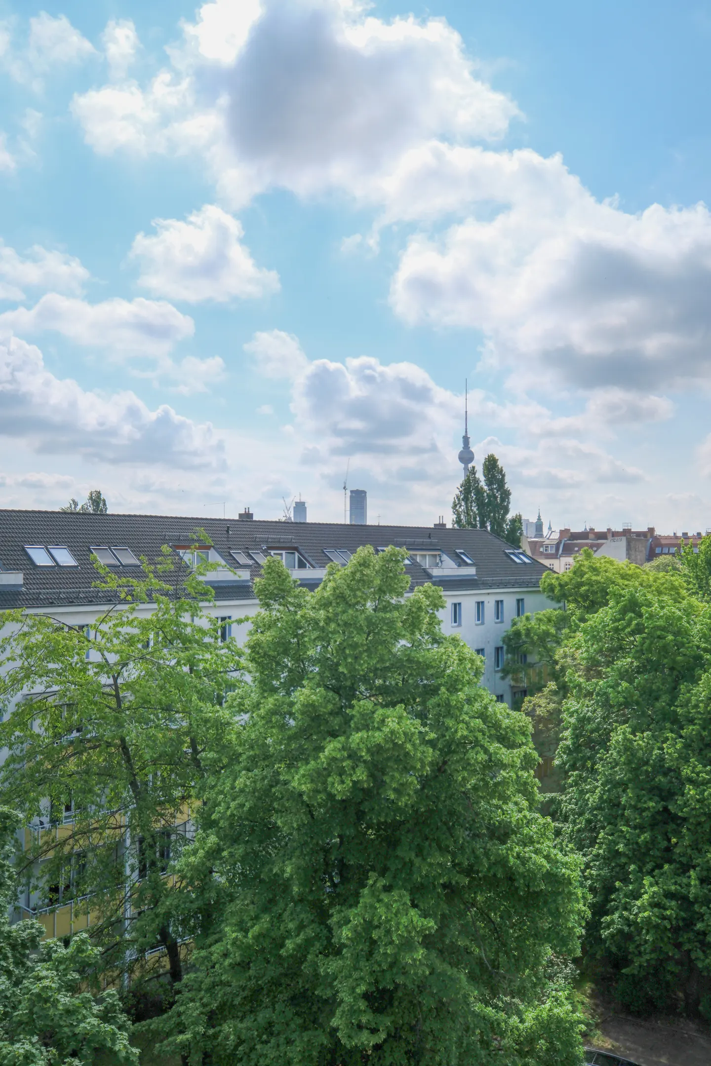 View of Berlin cityscape with green trees, white buildings, and the TV Tower under a cloudy blue sky.