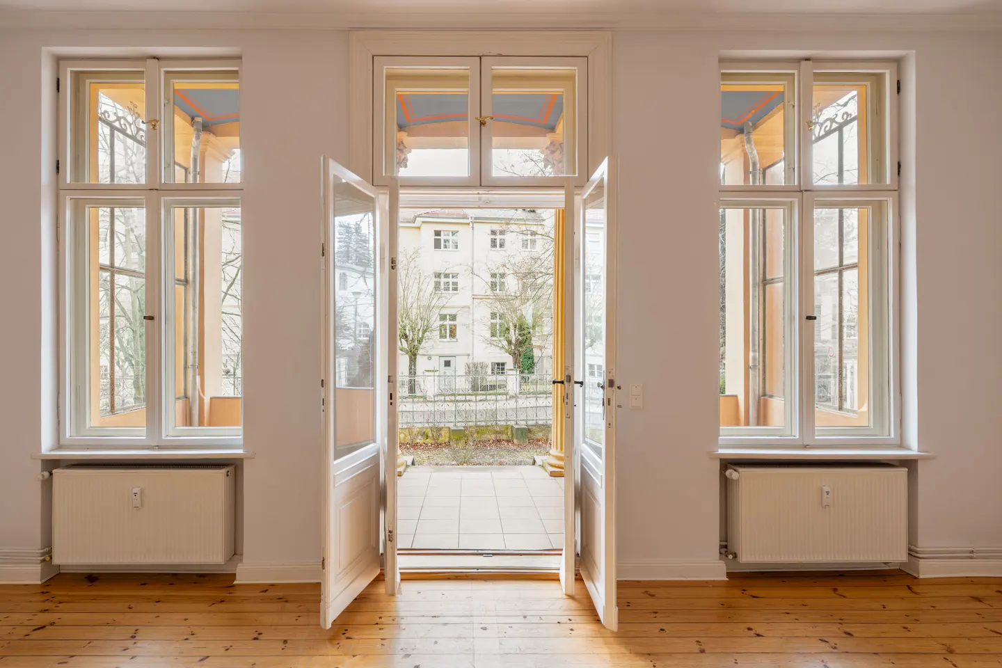Bright room with wood floor, white walls, and open double doors to a patio. Two large windows flank the doors.
