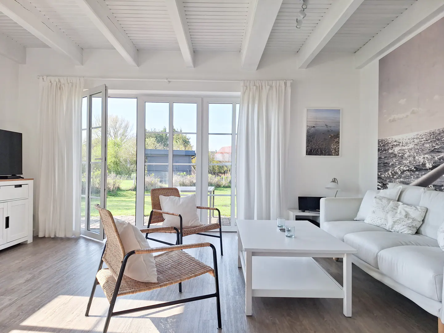 Bright living room with white walls, ceiling beams, and curtains. Wicker chairs face open glass doors to a green yard. A white sofa and table sit on wood floors.