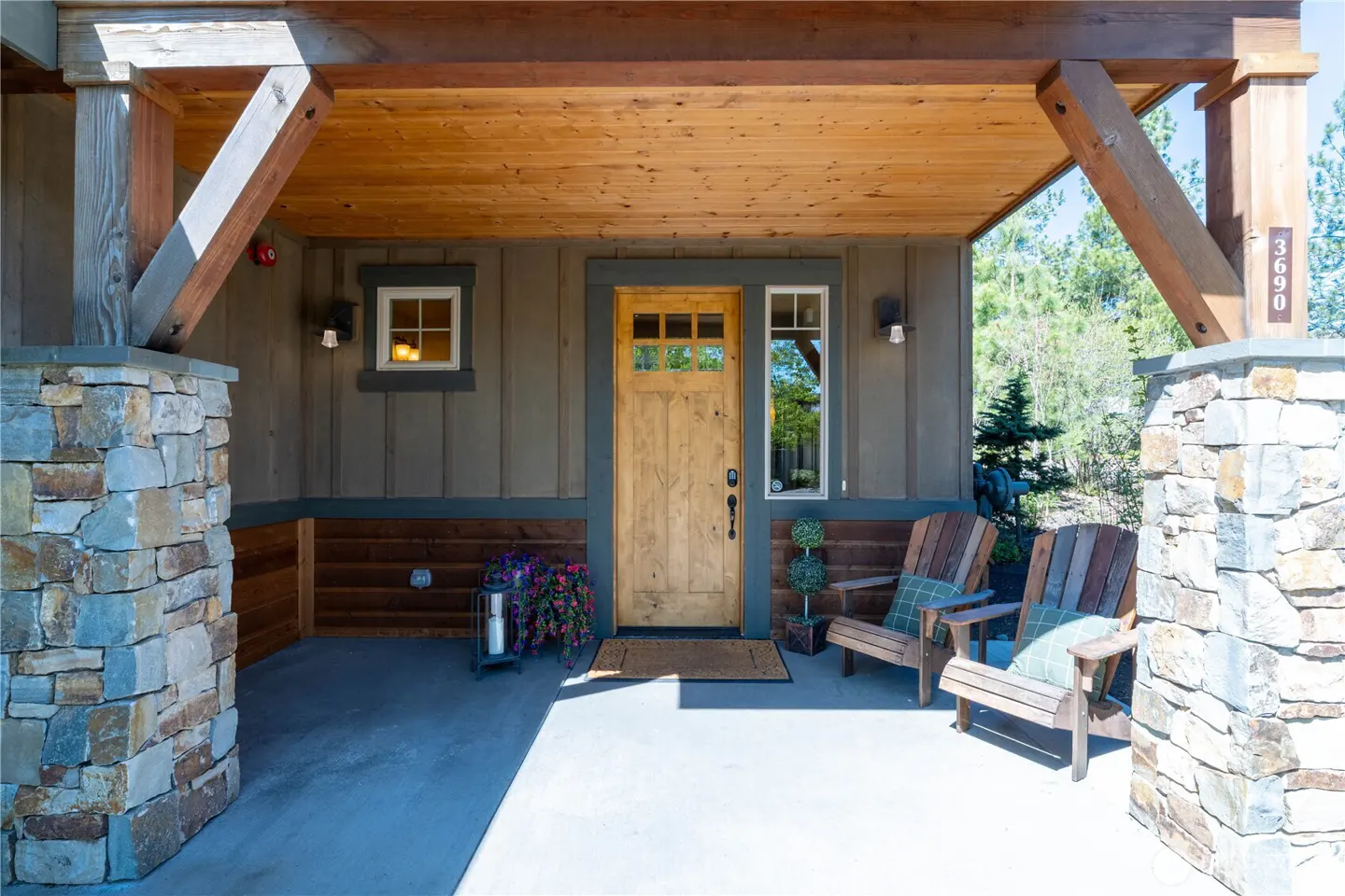 Rustic covered porch with stone pillars, wood siding, a wooden front door, and two wooden chairs with green cushions.