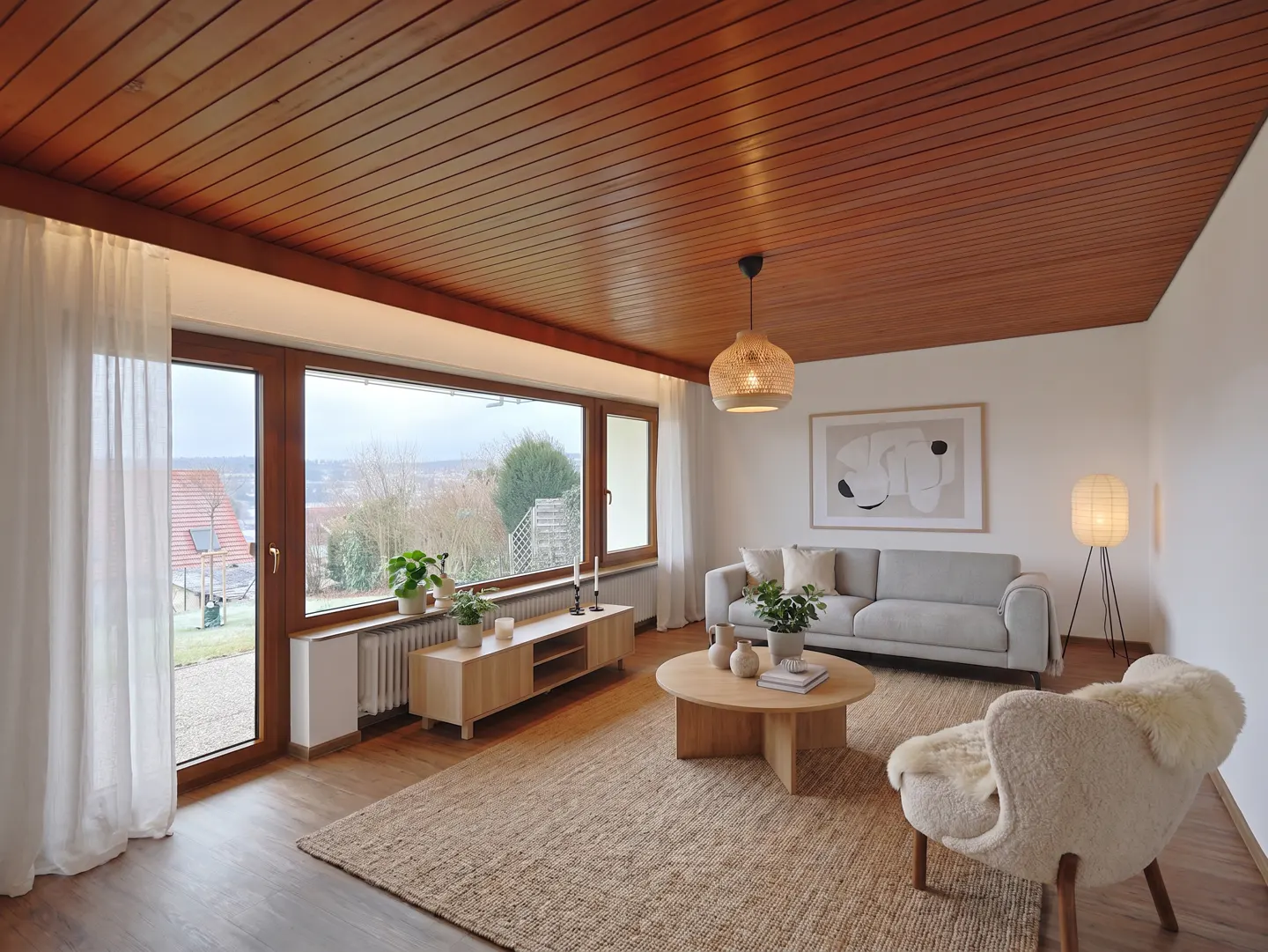 Bright living room with wood-paneled ceiling, large window, gray sofa, round coffee table, and a sheepskin armchair on a jute rug.