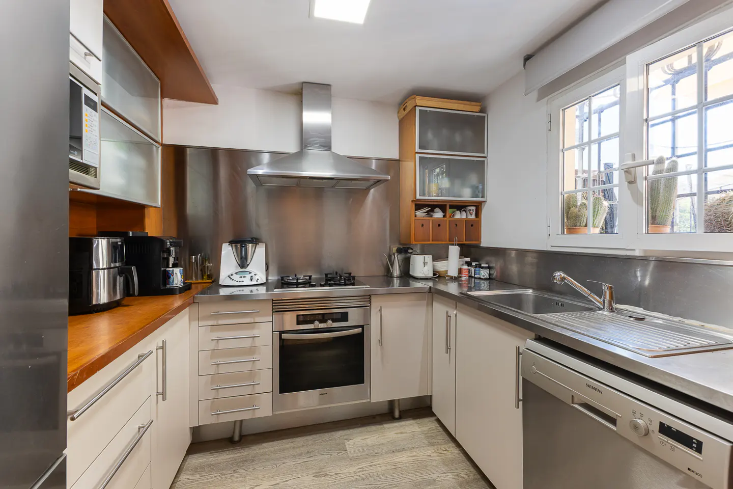 A modern kitchen with stainless steel backsplash, light cabinets, and wood countertops. A window with plants provides natural light.