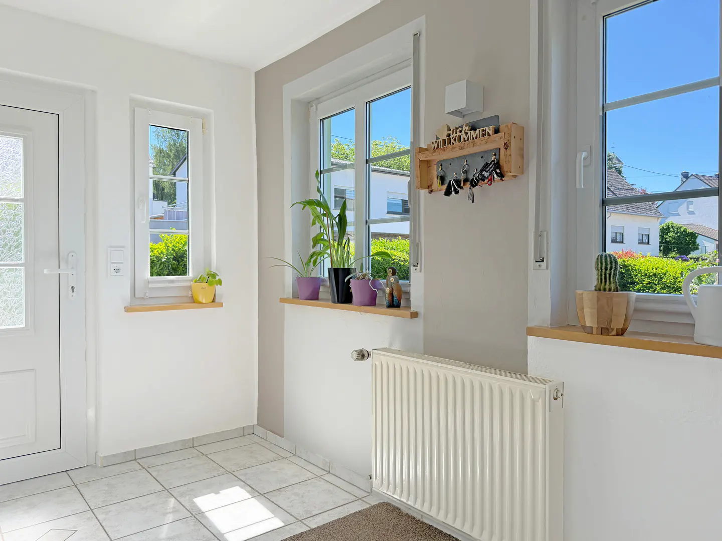 Bright entryway with white tiled floors, windows, a radiator, potted plants, and a wooden key rack on a beige wall.