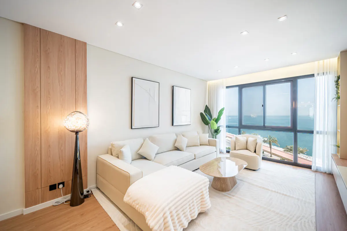 Modern living room with cream sectional sofa, round coffee table, floor lamp, and large window overlooking the ocean.