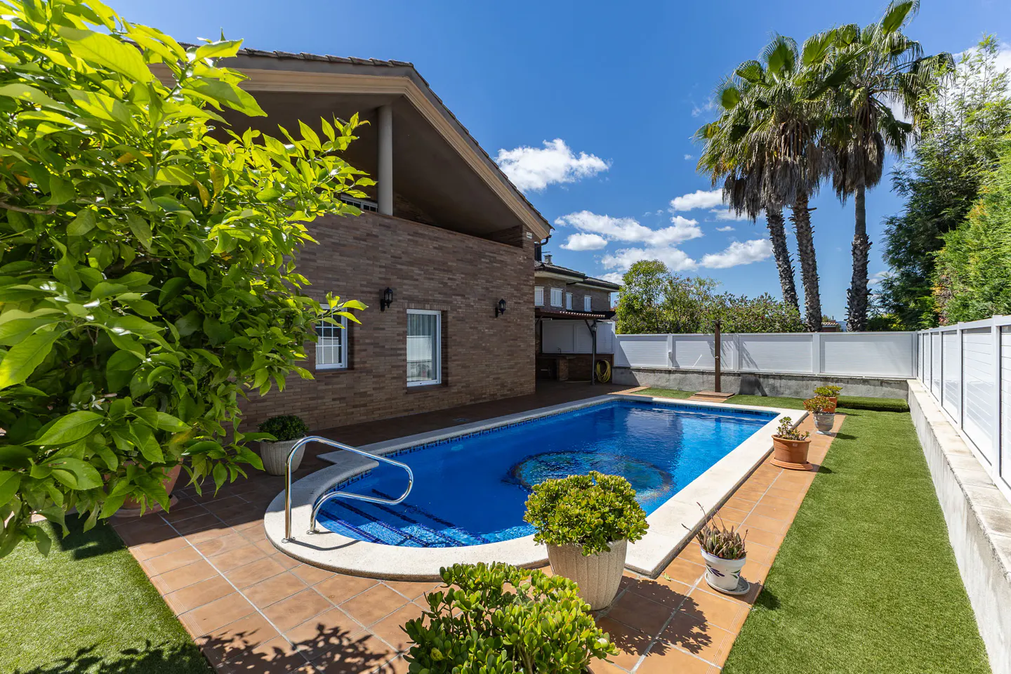 Backyard view of a rectangular blue swimming pool with a mosaic design, surrounded by green grass and a brick house.