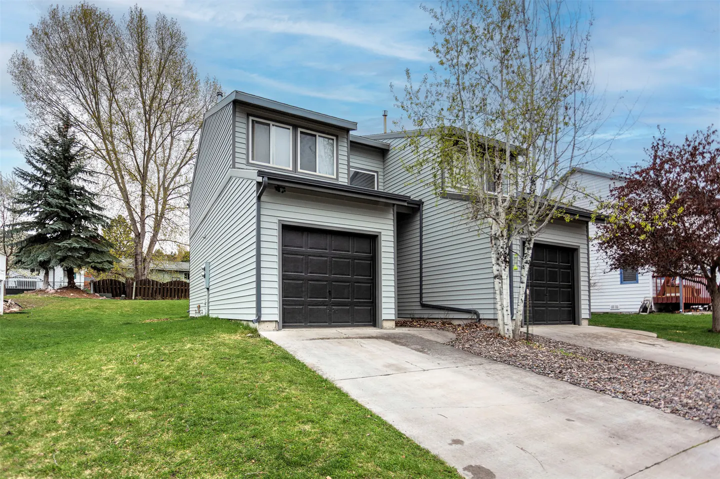 Two-story light blue townhouse with black garage doors, a concrete driveway, and a green lawn under a partly cloudy sky.