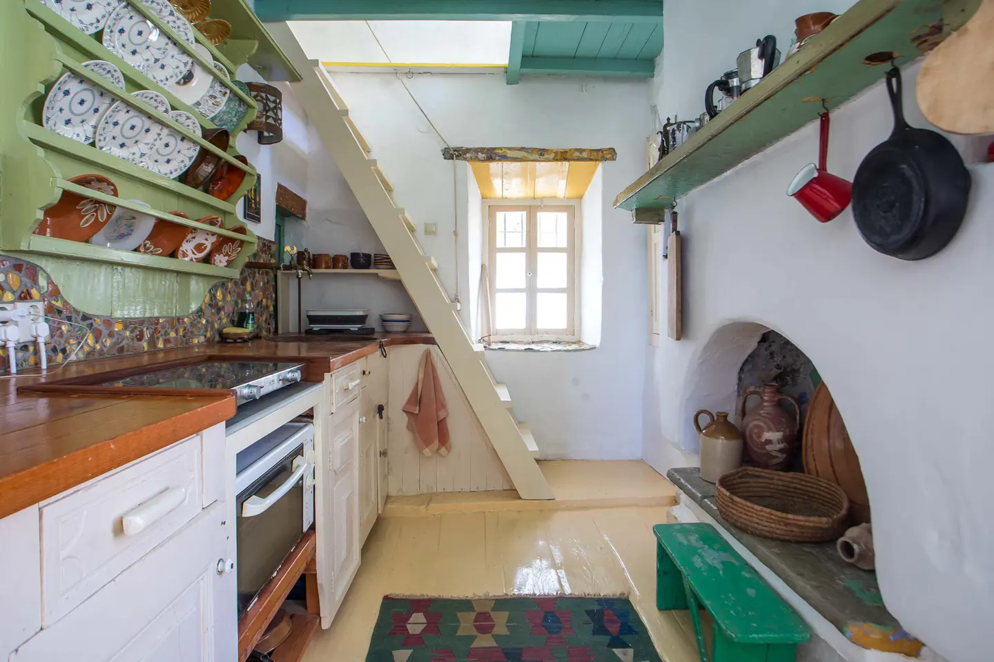 Rustic kitchen with white cabinets, green shelving, a wooden staircase, and a fireplace with hanging cookware.