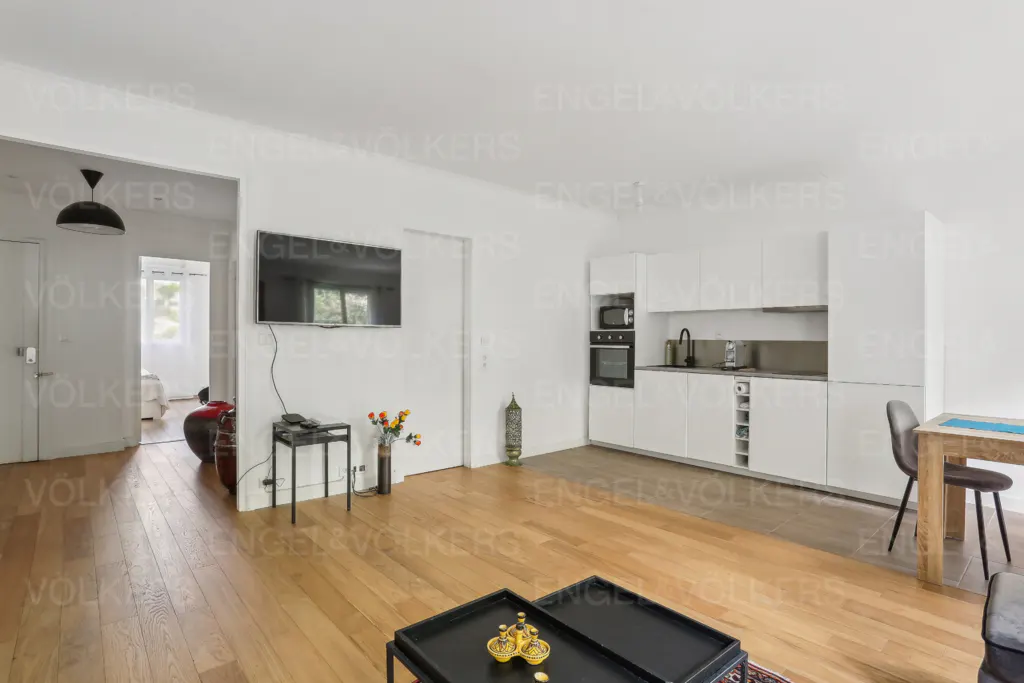 Bright, open-concept apartment with hardwood floors, white walls, and a modern kitchen. A TV hangs above a small table with flowers.