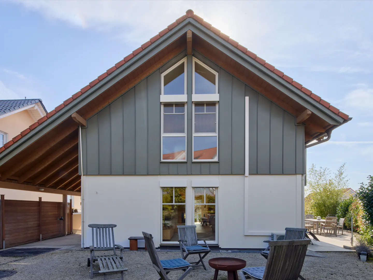 Exterior view of a two-story house with gray siding, white trim, and a red tile roof. A gravel patio has wooden chairs and a table.
