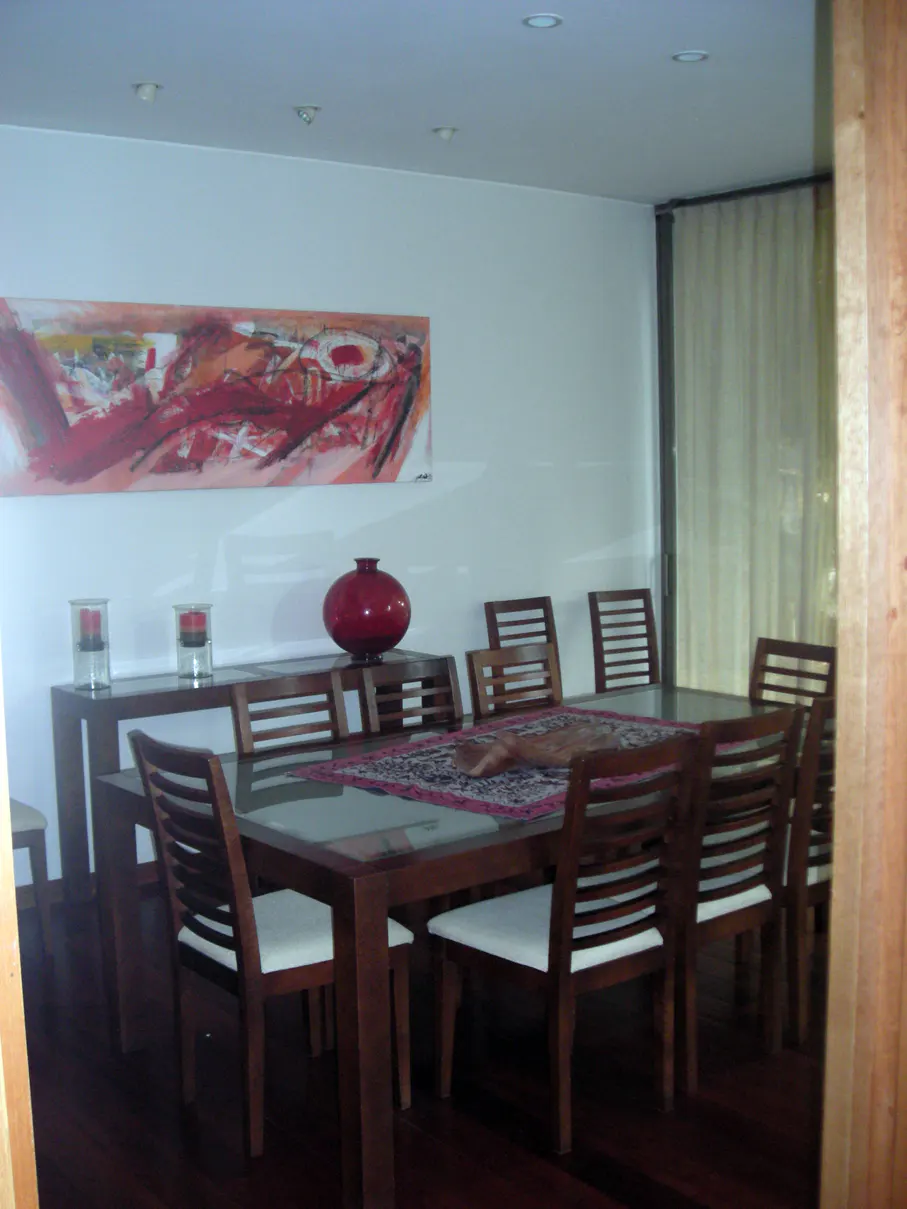 Dining room with a long wood table and eight chairs. A red vase sits on a console table under a red abstract painting.