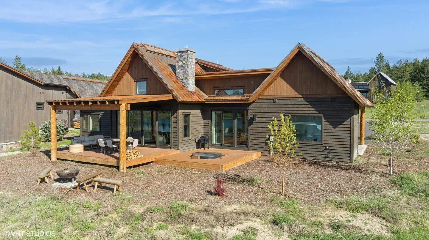 Exterior view of a brown, wood-sided house with a stone chimney, a wooden deck, and a fire pit with benches.