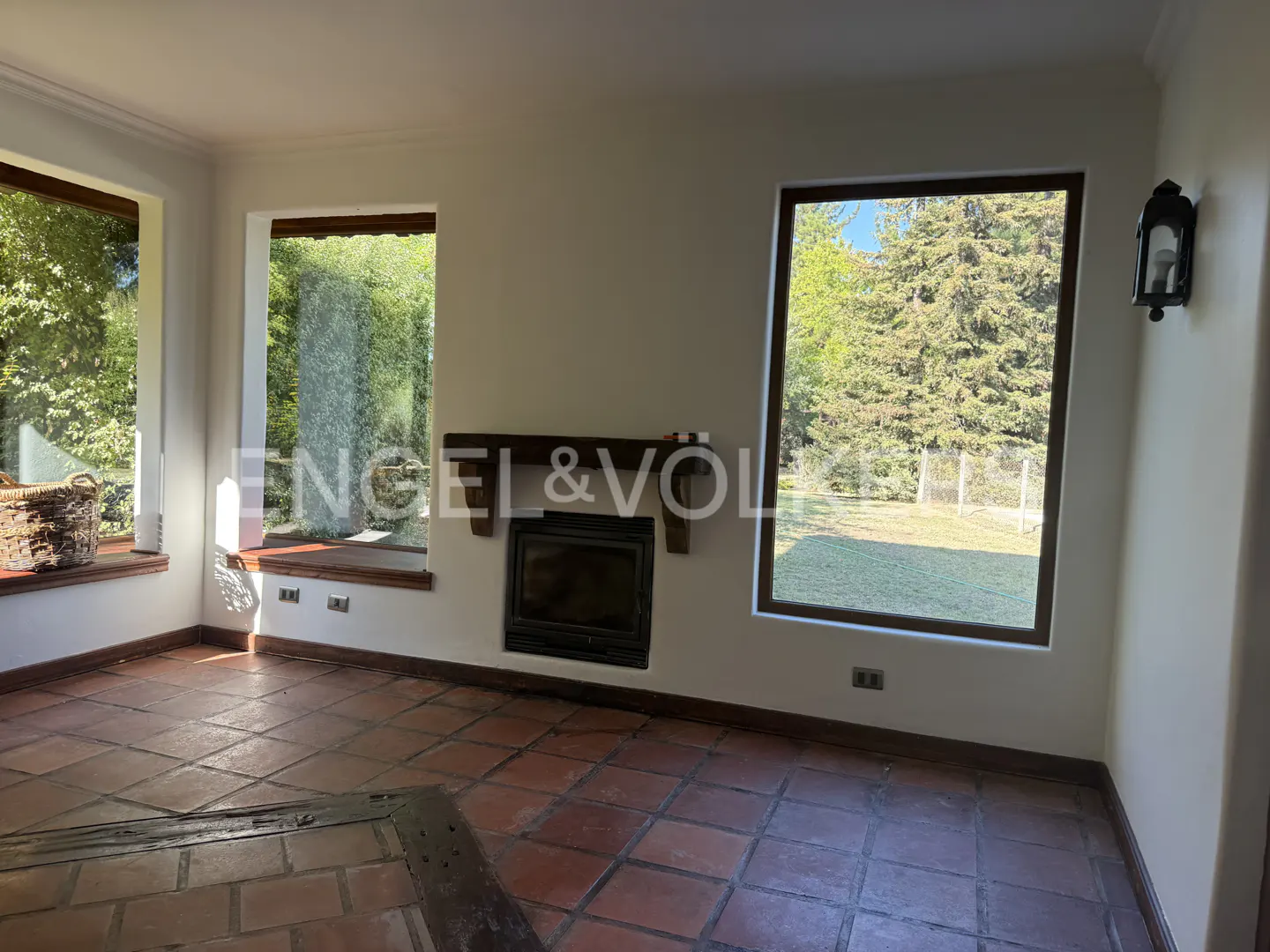 A living room with terracotta tile floors, a fireplace, and large windows overlooking a green lawn.