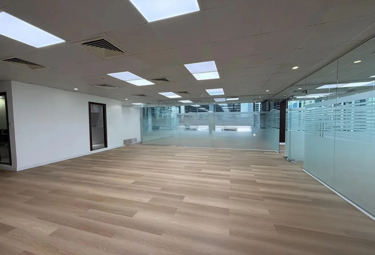 Empty office space with light wood floors, white walls, and glass-walled rooms with frosted stripes. Recessed lighting in the ceiling.