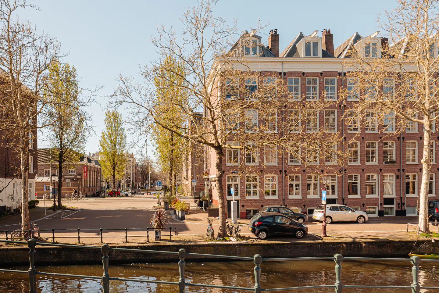 Amsterdam street view with a canal in the foreground. Brick buildings line the street with cars parked along the side.