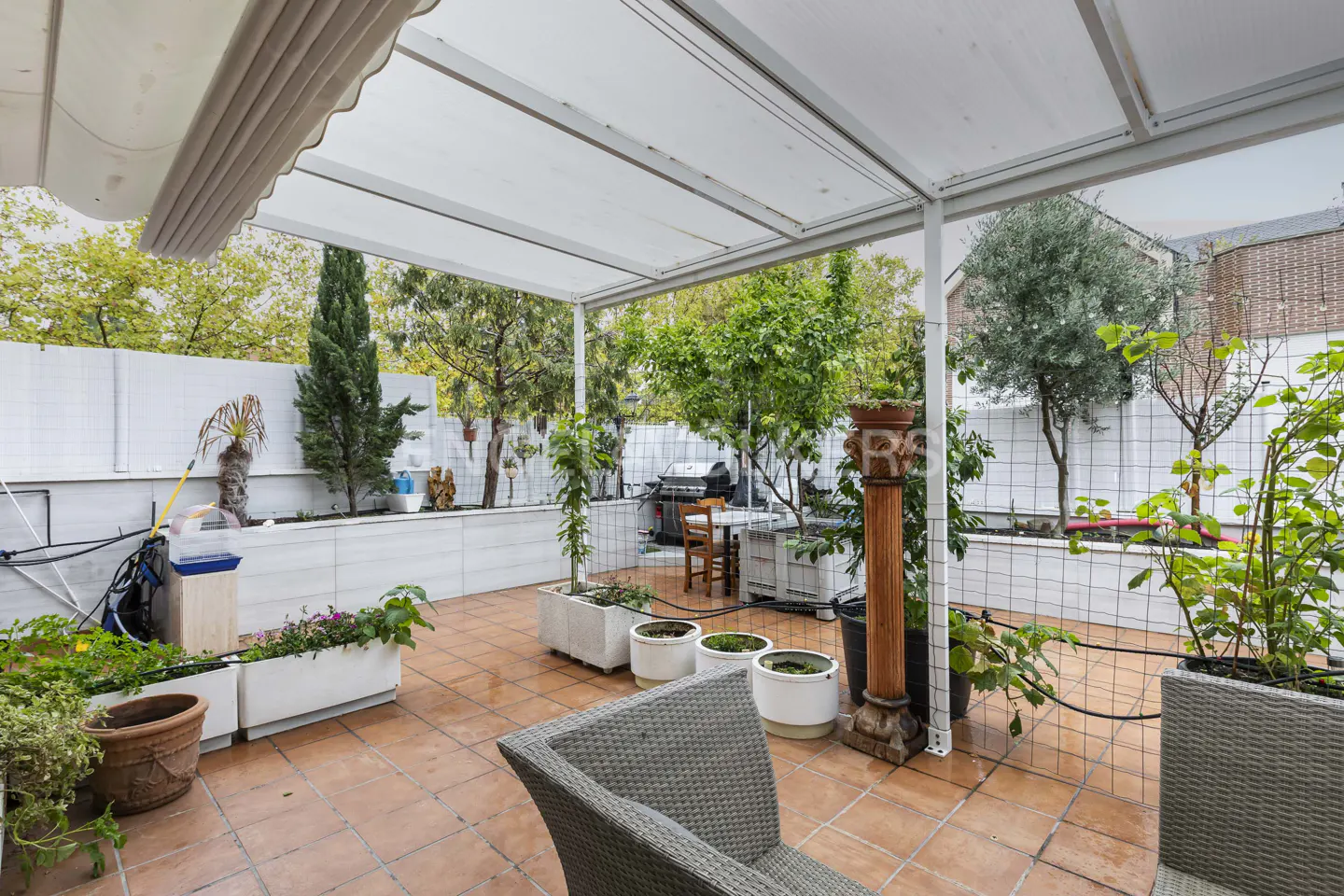 Covered patio with terracotta tiles, potted plants, gray wicker chairs, and a white fence against a backdrop of trees.