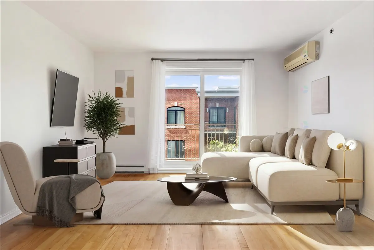 Bright living room with hardwood floors, white walls, and a large window. A beige sectional sofa and chair provide seating.