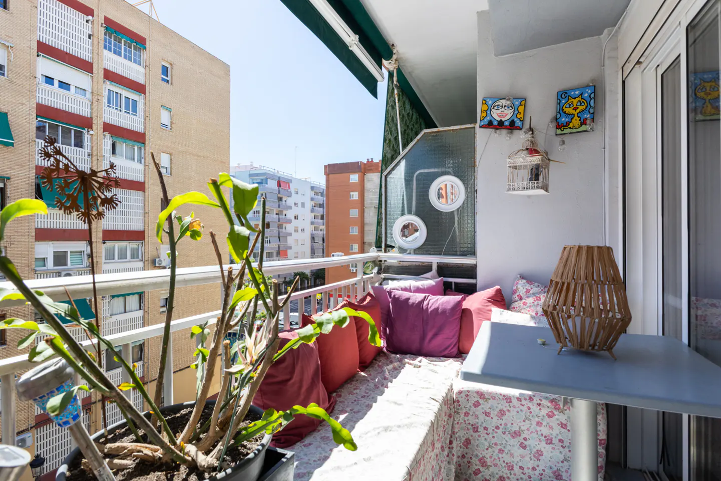 Cozy balcony with city view. Cushions line a bench, a small table holds a lamp, and plants add greenery. Art and a birdcage decorate the wall.