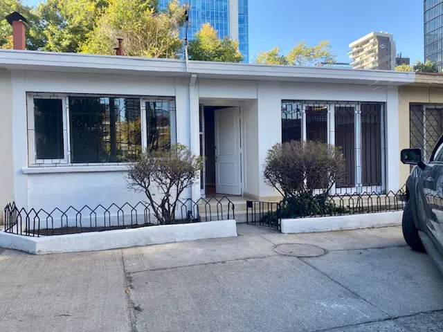 Exterior of a white, one-story house with an open front door, black window bars, and small front yard. Tall buildings are in the background.