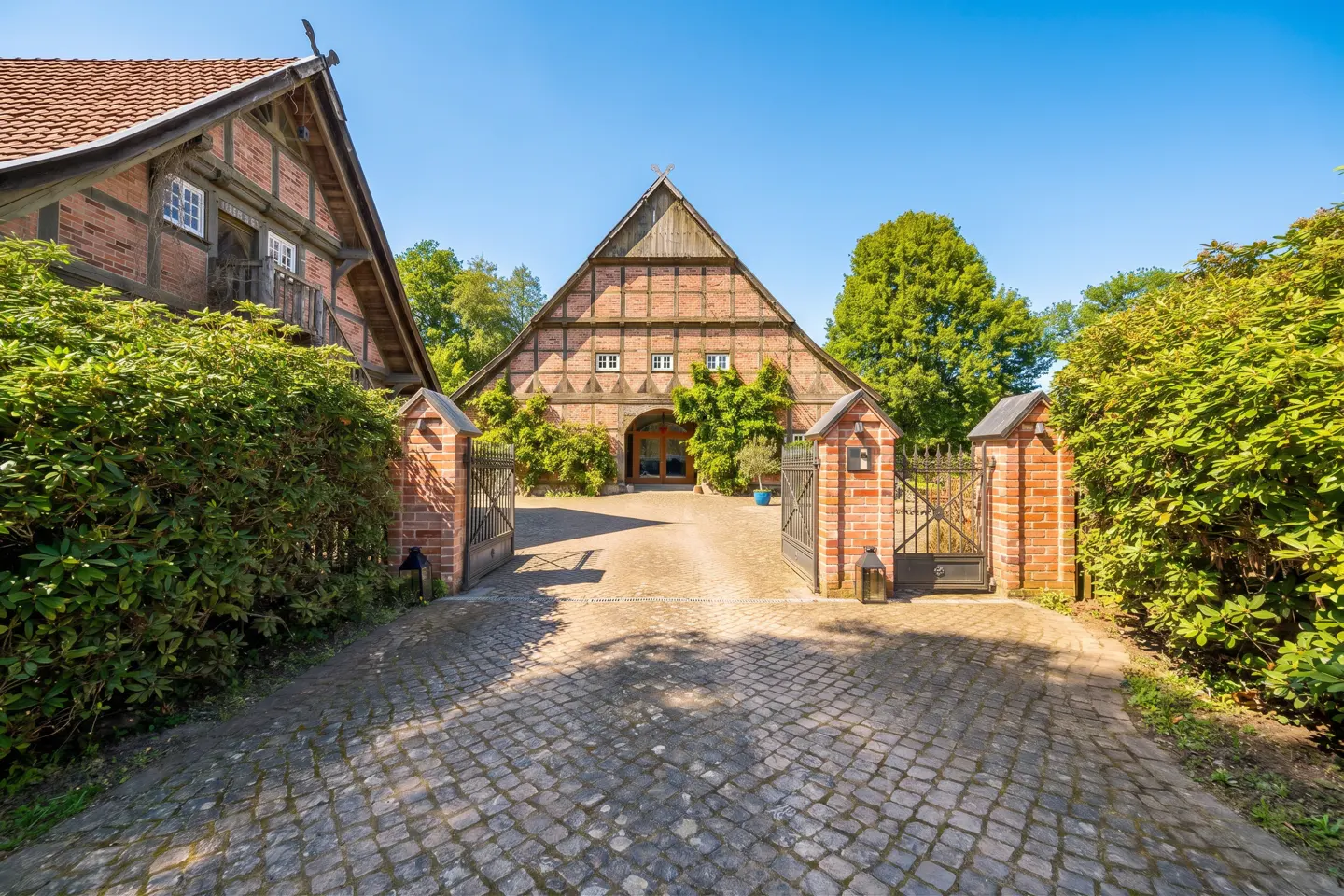 Cobblestone driveway leading to a traditional half-timbered brick farmhouse with a gated entrance and lush green bushes.