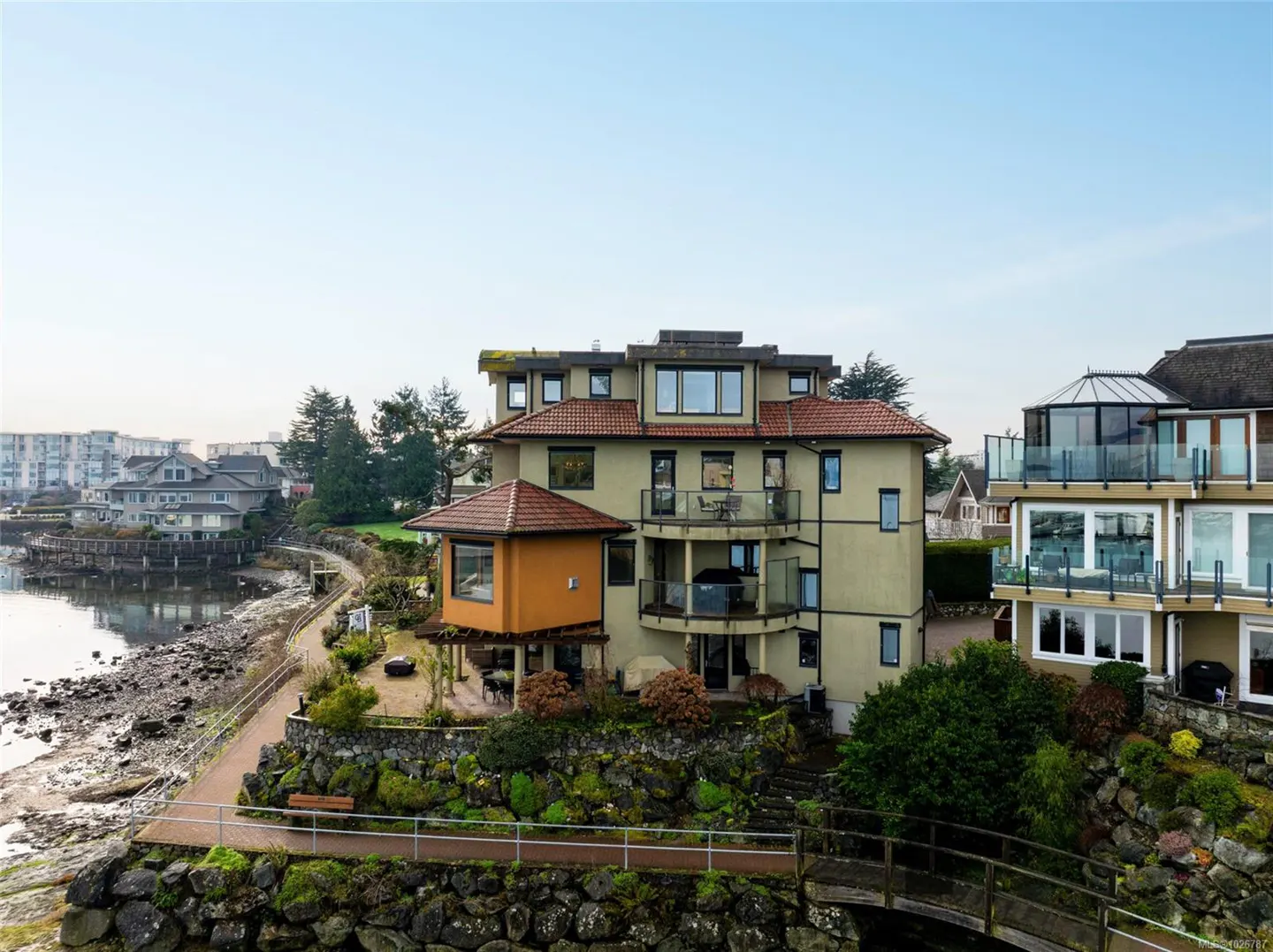 Multi-story tan building with red tile roof and balconies overlooking the ocean. A stone wall and path are in the foreground.