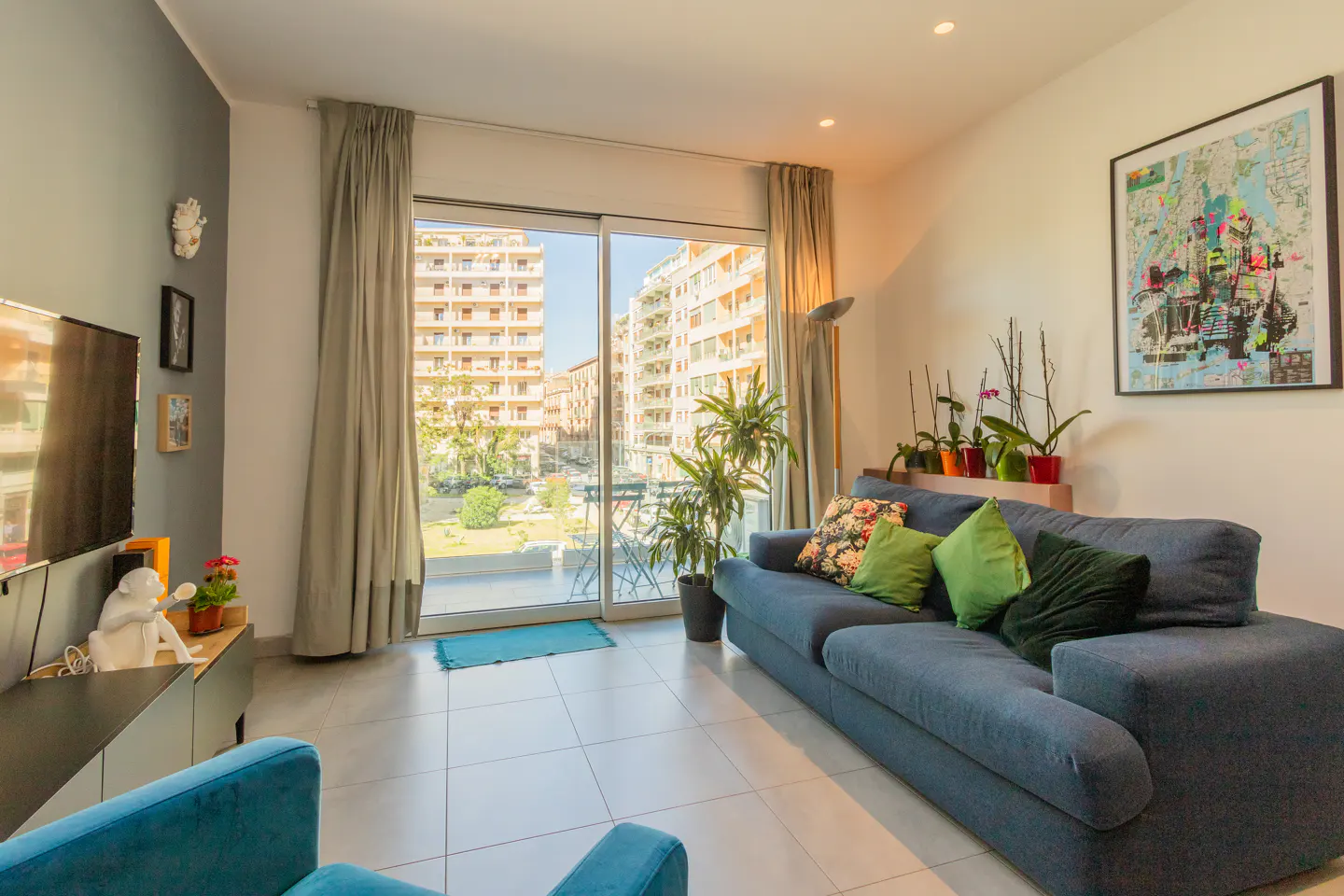 Bright living room with a blue sofa, green pillows, and a balcony view of buildings and greenery. A colorful map hangs on the wall.