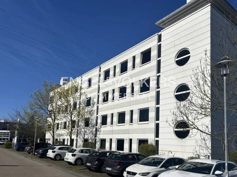 Exterior view of a modern, multi-story white office building with a row of parked cars in front on a sunny day.