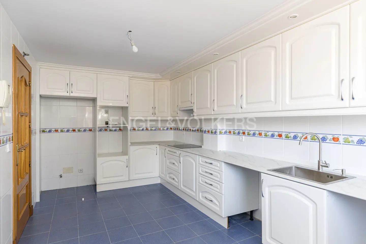 Bright kitchen with white cabinets, marble countertops, and blue tiled floor. A wooden door is on the left.
