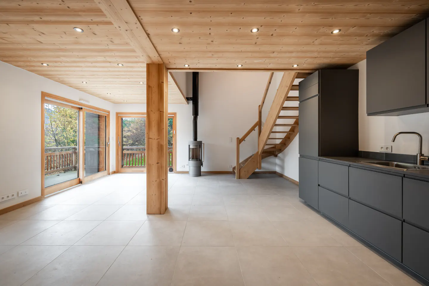Bright, modern interior with wood ceiling, gray cabinets, and wood staircase. Sliding glass doors lead to a balcony with a view of trees.